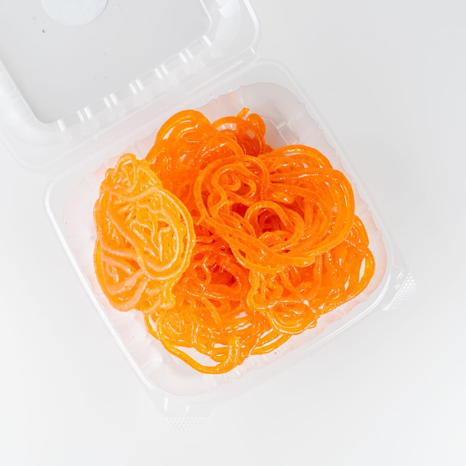 Bright orange jalebi sweets arranged in a clear takeaway container on a white surface.