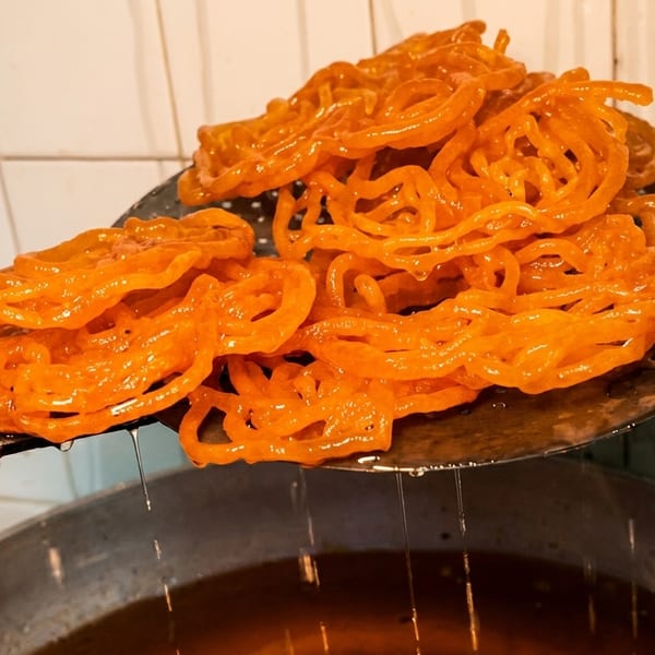 Golden-orange jalebi, a sweet Indian dessert, being lifted from a pot of syrup with a slotted spoon.