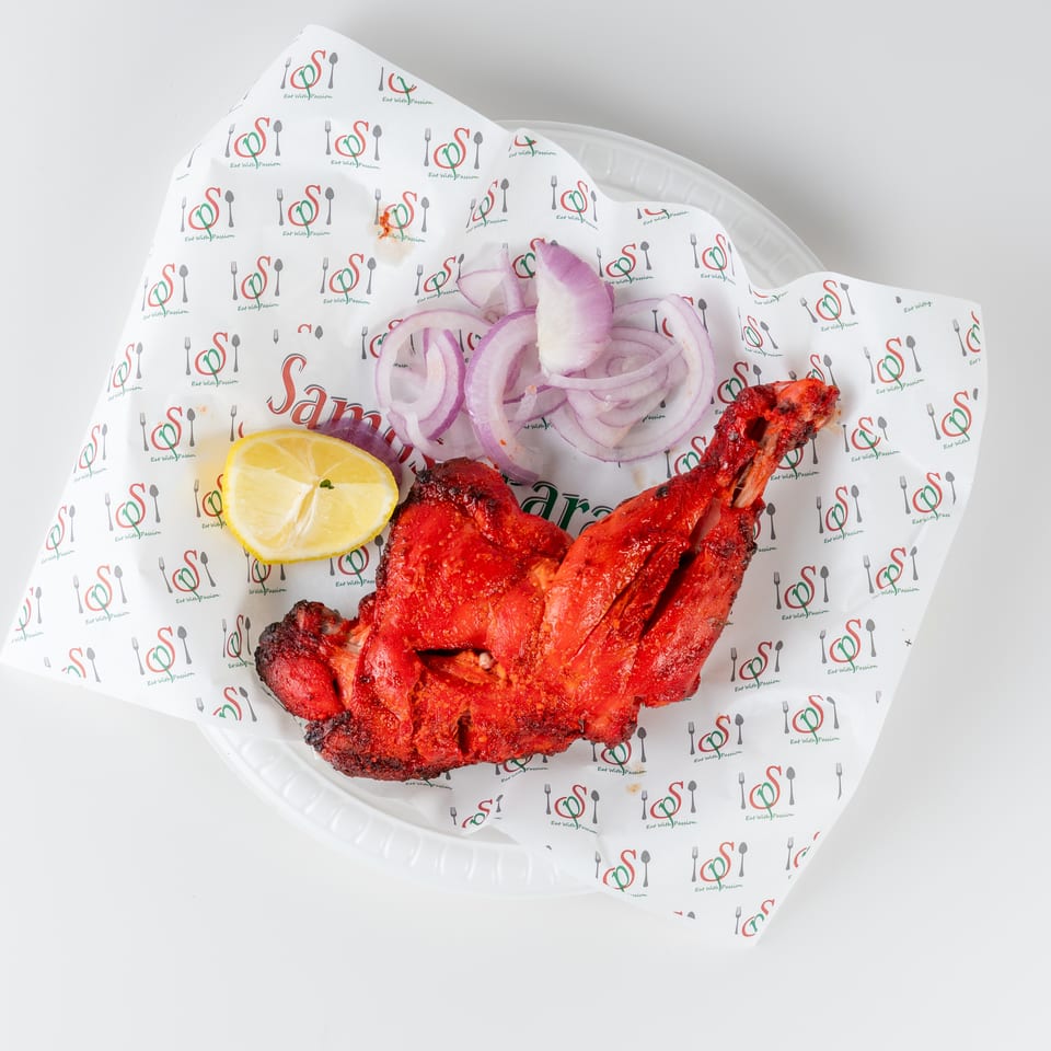 Red tandoori chicken pieces served with sliced red onion and lemon on a branded paper-lined plate.