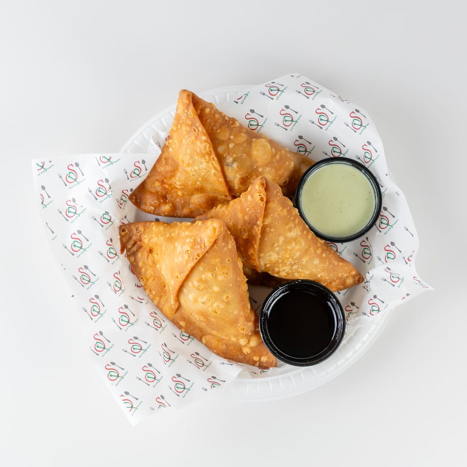Golden fried samosas arranged on a branded paper-lined tray with green and dark dipping sauces.